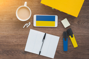 flag of Ukraine, smartphone, wireless headphones, textbook, notepad on a working wooden brown...