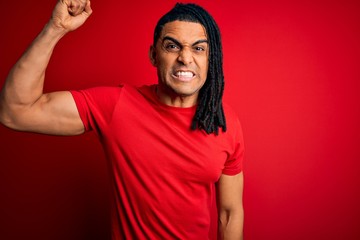 Young handsome african american afro man with dreadlocks wearing red casual t-shirt angry and mad raising fist frustrated and furious while shouting with anger. Rage and aggressive concept.