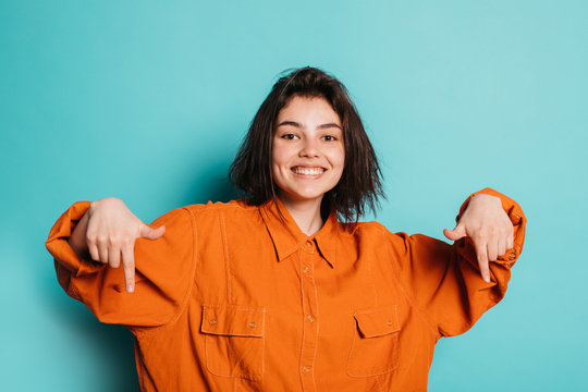 Picture of positive cheerful young woman or teenager isolated over blue background. Happy girl point down with index fingers. Stylish modern woman wear orange shirt.