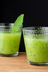 Top view of two glasses with cucumber juice, one with mint leaf, with selective focus, on wooden box, black background, vertical