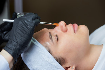 Cosmetologist wearing black disposable medical gloves uses a brush to apply a superficial transparent face peeling to a young woman. A cosmetology procedure in a beauty salon for skin cleaning.