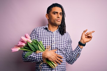 Young african american afro romantic man with dreadlocks holding bouquet of pink tulips Pointing with hand finger to the side showing advertisement, serious and calm face