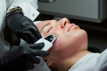 A close-up photo of the woman's face after a warming lotion during an ultrasonic procedure in the cosmetology office. A cosmetologist in disposable gloves peeling face with an ultrasonic skin scrubber