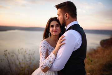 Obraz premium The bride and groom are hugging on the background of the lake during sunset.