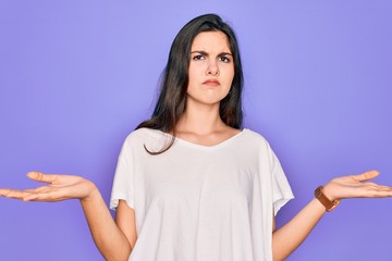 Young beautiful brunette woman wearing casual white t-shirt over purple background clueless and confused with open arms, no idea concept.