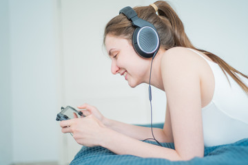 Joyful girl lies on the bed and plays the game console in headphones. Portable play in apartment