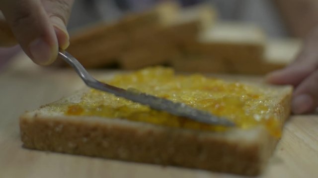 Close up people's hand spreading organic orange marmalade on sliced wholewheat bread with a knife on a wooden table. Healthy eating concept.