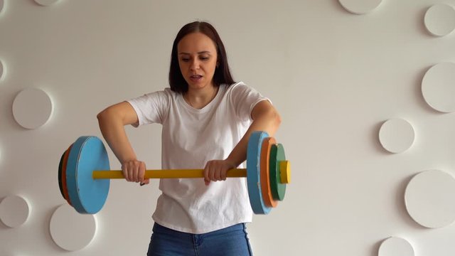 Young woman lifts multicolored barbell against white patterned wall. Adult lady doing sport, using all her strength.