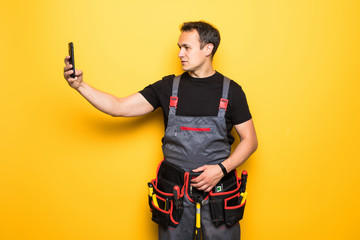 Young man worker with tools belt holding phone isolated on yellow background