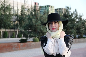 a beautiful woman in a black jacket, black hat over a beige scarf, walk around the city alone, close-up portrait. without gloves, her hands are pressed to her chest