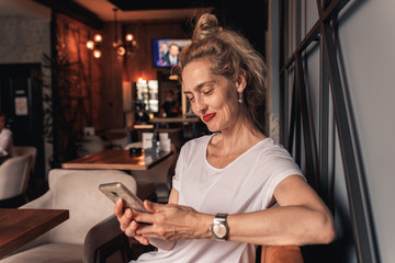 Fototapeta na wymiar Smiling senior woman sitting in cafe drinking coffee and using smartphone.