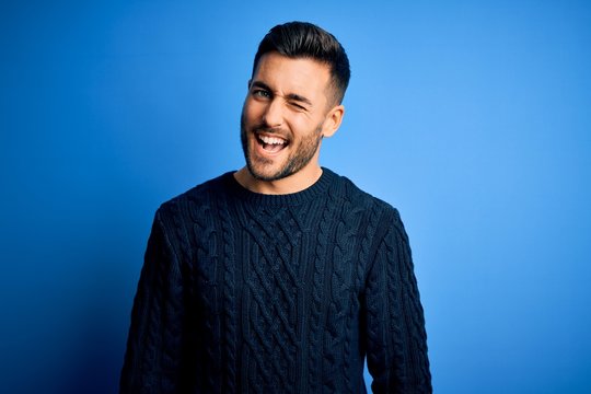 Young handsome man wearing casual sweater standing over isolated blue background winking looking at the camera with sexy expression, cheerful and happy face.