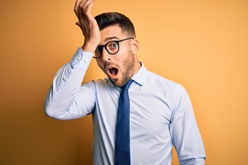 Young handsome businessman wearing tie and glasses standing over yellow background surprised with...