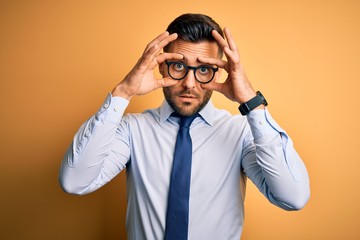 Young handsome businessman wearing tie and glasses standing over yellow background Trying to open eyes with fingers, sleepy and tired for morning fatigue