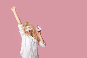 Relaxed young woman in pajamas and sleep masks on a pink background. Alarm clock woke up the girl