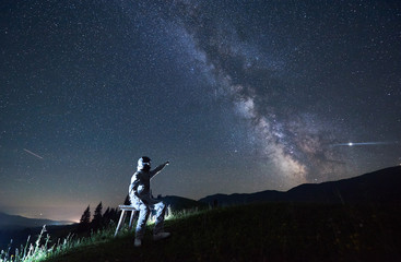 Illuminated man in white space suit and helmet sitting on stool on a hill, pointing at Milky Way, bright Polar Star and shooting stars in the starry sky at night. Concept of space travel, cosmonautics