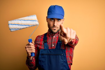 Young handsome painter man painting wall using roller over isolated yellow background pointing with...