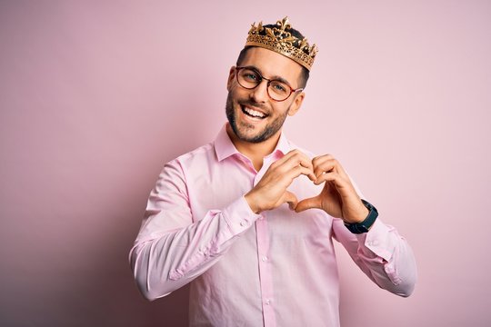 Young handsome business man wearing golden crown as king over pink background smiling in love showing heart symbol and shape with hands. Romantic concept.