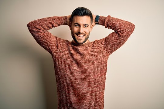 Young handsome man wearing casual sweater standing over isolated white background relaxing and stretching, arms and hands behind head and neck smiling happy