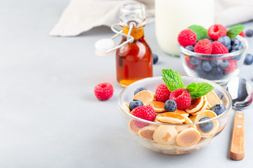 Tiny cereal pancakes served with maple syrup, raspberry, blueberry and milk, in glass bowl, horizontal, copy space