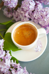 Fresh espresso in a white cup and lilac flowers on the background.