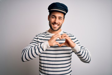 Young handsome sailor man with beard wearing navy striped uniform and captain hat smiling in love showing heart symbol and shape with hands. Romantic concept.