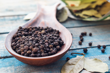Black pepper in a large wooden spoon on a background of blue boards near a bay leaf.