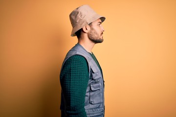 Handsome tourist man with beard on vacation wearing explorer hat over yellow background looking to side, relax profile pose with natural face and confident smile.