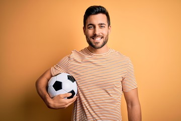 Fototapeta Handsome player man with beard playing soccer holding footballl ball over yellow background with a happy and cool smile on face. Lucky person. obraz