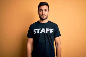 Young handsome worker man with beard wearing staff uniform t-shirt over yellow background with a confident expression on smart face thinking serious
