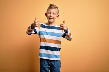 Young little caucasian kid with blue eyes wearing colorful striped shirt over yellow background success sign doing positive gesture with hand, thumbs up smiling and happy. Cheerful expression