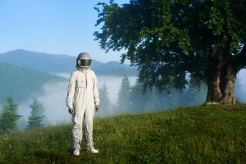 Portrait of a spaceman wearing white space suit and a helmet standing on a green mountain meadow on a hill, big old tree and fantastic foggy spruce forest in a valley on background. Travel concept © anatoliy_gleb