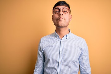 Young handsome hispanic business man wearing nerd glasses over yellow background making fish face...