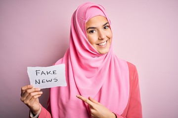 Young beautiful girl wearing muslim hijab holding paper with fake news message very happy pointing with hand and finger