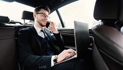 Young businessman talking on phone in car using laptop