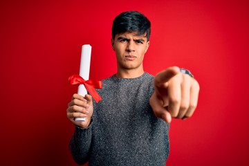 Young student man holding university graduated diploma degree over red background pointing with...