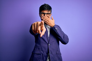 Young handsome business man wearing jacket and glasses over isolated purple background laughing at you, pointing finger to the camera with hand over mouth, shame expression