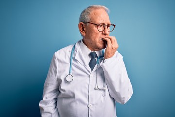 Senior grey haired doctor man wearing stethoscope and medical coat over blue background looking stressed and nervous with hands on mouth biting nails. Anxiety problem.