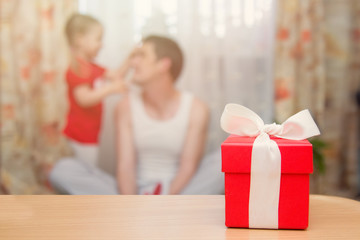 A red gift box with a white ribbon stands on a table in the background father with a small daughter. Father's Day Holiday Concept