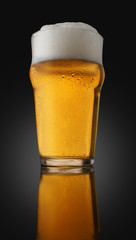 A pint of beer with foam in a glass with water drops on a dark background with gradient lighting and reflection in the surface