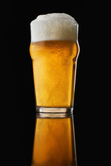 A pint of beer with foam in a glass with water drops on a dark background and reflection in the surface