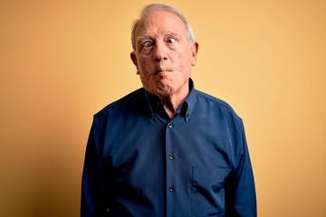 Grey haired senior man wearing casual blue shirt standing over yellow background making fish face with lips, crazy and comical gesture. Funny expression.
