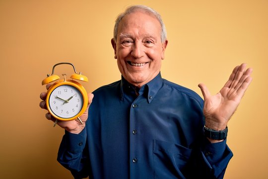 Senior grey haired man holding vintage alarm clock over yellow background very happy and excited, winner expression celebrating victory screaming with big smile and raised hands