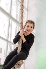 Obraz premium Schoolgirl doing physical exercises in the gym. Little happy girl is trying to climb up the old gymnastic rope. Portrait of a cheerful child who goes in for sports. Vertical photo