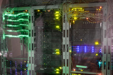Powerful computers work at the data center racks behind a glass wall. Bright indication of equipment in the server room. Computer hardware is in a dark room. Technological concept.