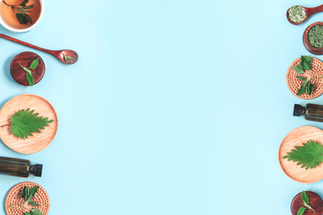 Frame of fresh young spring leaves, dry nettle, salt with herbs in wood spoon,bottle on pastel blue background. Apothecary, healthy food or herbal cosmetics concept. Flat lay, copy space