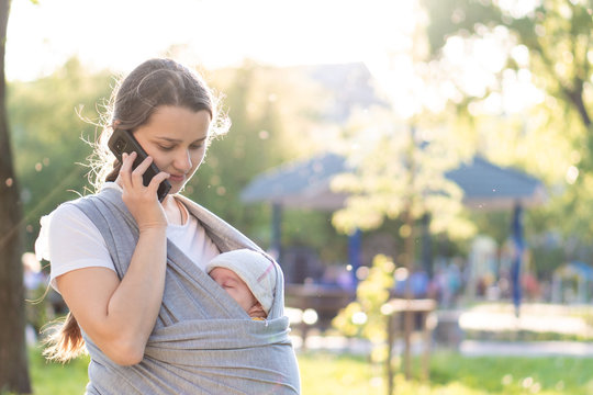 Motherhood, care, infants, summer, communication, parenting concept - Young beautiful mom with a newborn baby in a sling walks and talks on the phone in the backlight of a sunset in the park.