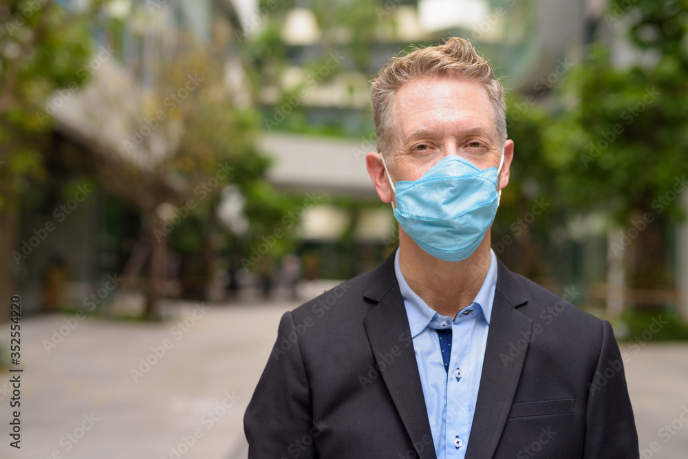 Wall mural Face of mature businessman with mask for protection from corona virus outbreak with nature in the city