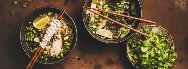 Asian cuisine lunch. Flat-lay of Vietnamese rice noodle chicken soup Pho Ga with cilantro, soy sprouts, fresh greens, lime in bowls with chopsticks over dark background, top view, wide composition