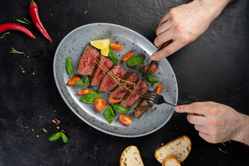 Man cuts grilled beef roast beef with vegetables and herbs with a table knife and fork.
